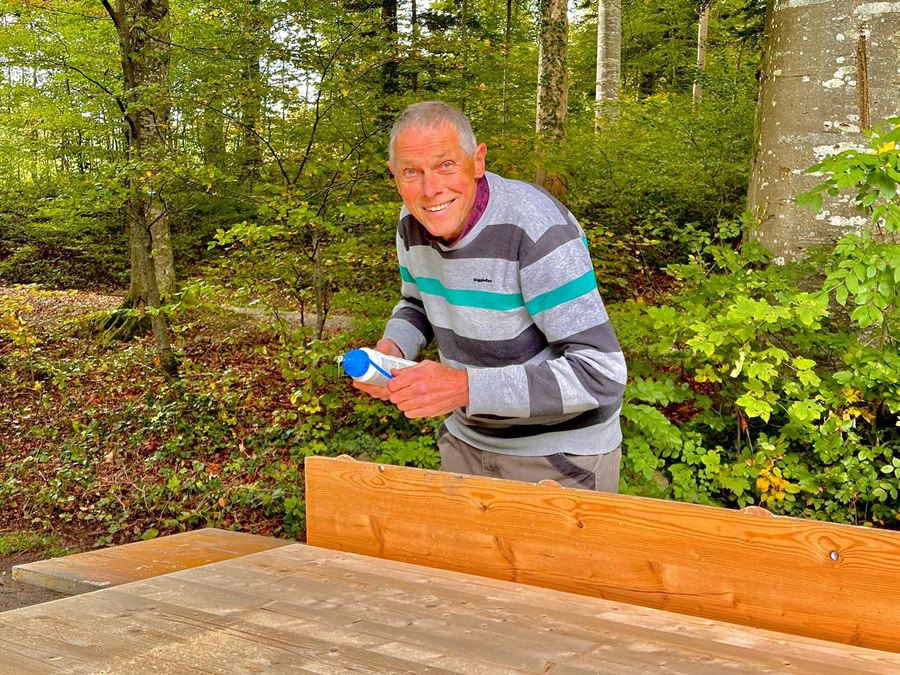 Ein Helfer repariert lachend eine Sitzbank im Wald